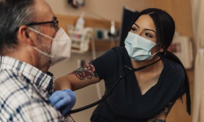 A PeaceHealth caregiver in a facemask covering uses a stethoscope to listen to a patient's lungs in a clinical setting.