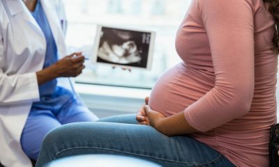 Photo of a pregnant woman with her hands on her belly in the foreground and her doctor with an ultrasound on a digital tablet in the background.