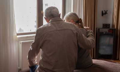 An older couple faces away from the camera to look out their bedroom window