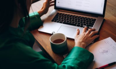 Woman at wooden table in green top viewing laptop screen. A green coffee cup and notepad are on the table next to her hands.