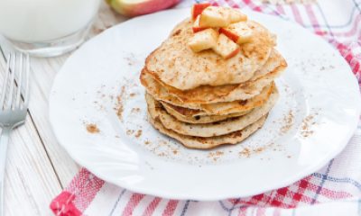 White plate with a stack of apple cinnamon pancakes. There is also a cup of milk and a fork on the table.