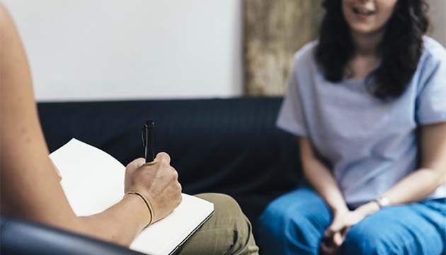 A person takes notes while another in the background speaks