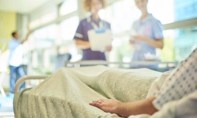 2 Healthcare providers with charts stand in the background, while a patient lays on a bed in the foreground