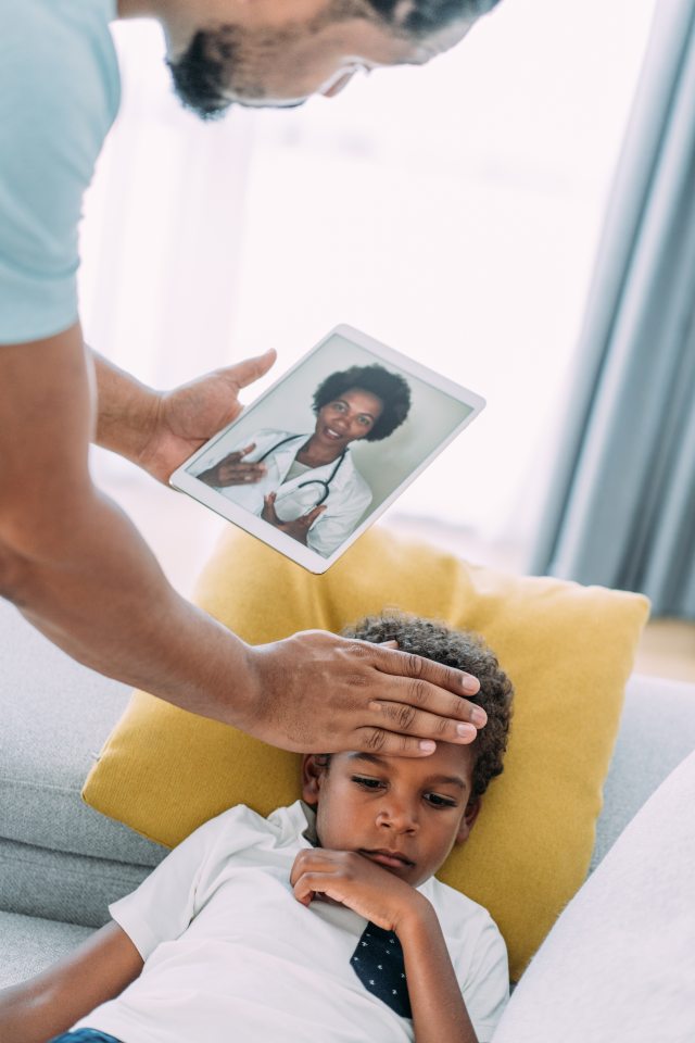 Man feels childs head for temperature while talking to doctor on mobile tablet