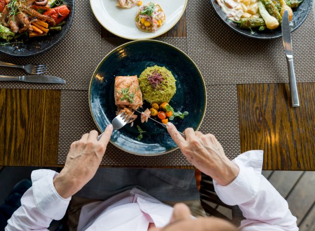 Birds eye view of a dinner plate with salmon, grains and vegetables with a person holding knife and fork.