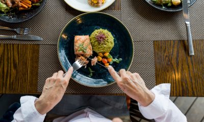 Birds eye view of a dinner plate with salmon, grains and vegetables with a person holding knife and fork.