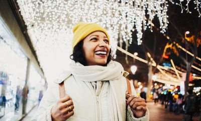 Woman in yellow hat and white scarf smiles outside brightly lit stores